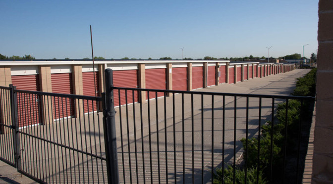 A view of gated entrance to a long row of outdoor storage units with red doors